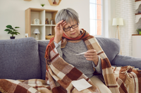 sick older woman checking temperature while wrapped in blanket on couch