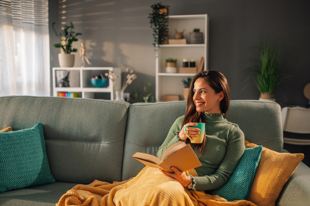 woman reading book on couch under blanket with cup of tea