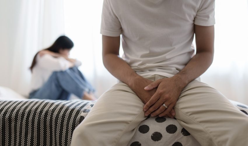 frustrated couple sitting on bed and man has hands covering lap