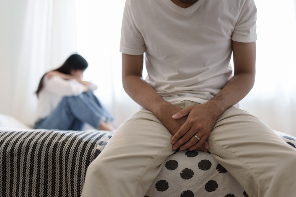 frustrated couple sitting on bed and man has hands covering lap