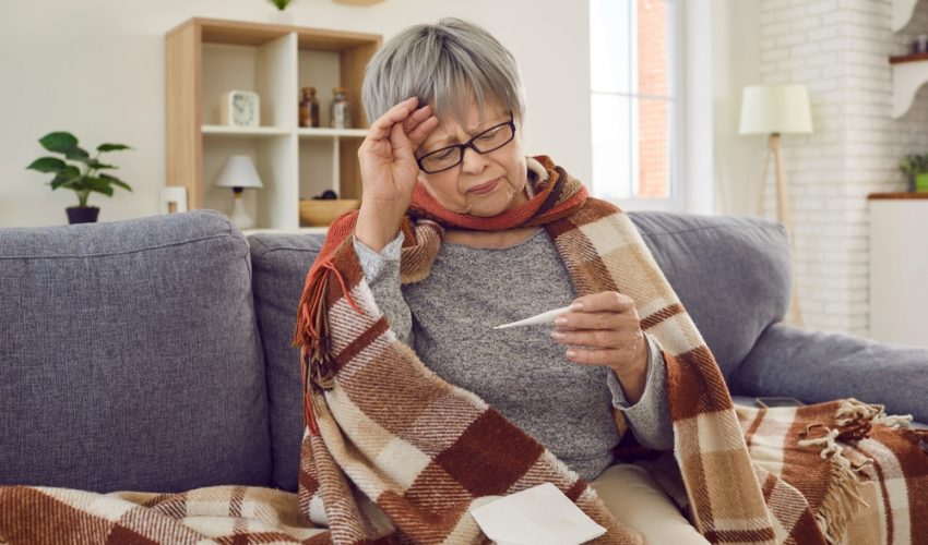 sick older woman checking temperature while wrapped in blanket on couch